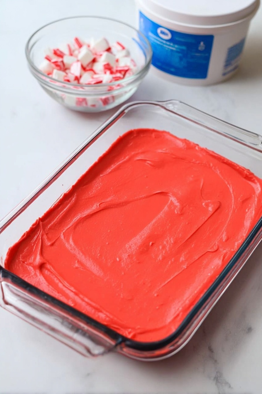 A slice of dessert with three visible layers sits on a white plate with a white marbled background. The bottom layer is a dark, crumbly chocolate crust with rough texture. The middle layer is thick and white, smooth and creamy in texture. The top layer is white whipped cream with many small and larger red candy pieces sprinkled over it. A silver fork is holding a piece of the dessert, showing all three layers clearly. The scene also shows a woman's hand holding the fork. photo taken with an iphone --ar 2:3 --v 7