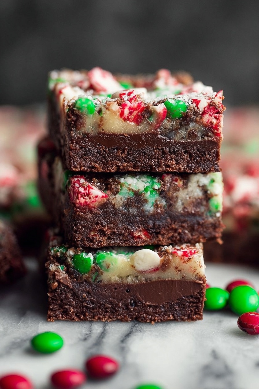 The image shows a stack of three thick, square-shaped brownies on a white marbled surface. Each brownie has two layers: the bottom layer is a dark brown, smooth and dense chocolate base, while the top layer is lighter with a mixture of red, green, and white candy pieces scattered evenly, adding a rough texture and colorful look. There are extra green and red candy pieces spread around the base of the stack. The top layer looks slightly melted around the candies, with a mix of colors blending subtly in places. Photo taken with an iphone --ar 2:3 --v 7