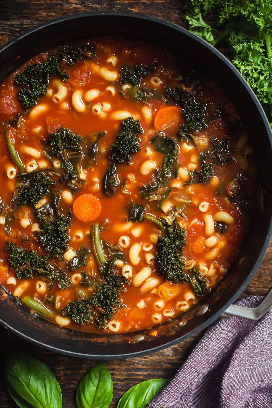 A white bowl with a blue scalloped edge holds a soup made of three main layers: the base layer is a clear red tomato broth, the middle layer is filled with small curved elbow macaroni pasta and white beans, and the top layer is a mix of dark green leafy kale pieces and bright green beans and carrots. On top, there are three thin slices of pale yellow cheese and several fresh bright green basil leaves scattered around. The bowl is placed on a brown wooden table with green basil leaves artfully scattered nearby, along with part of a white bowl with more soup and a black pot with a beige woven cover. The photo was taken with an iphone --ar 2:3 --v 7