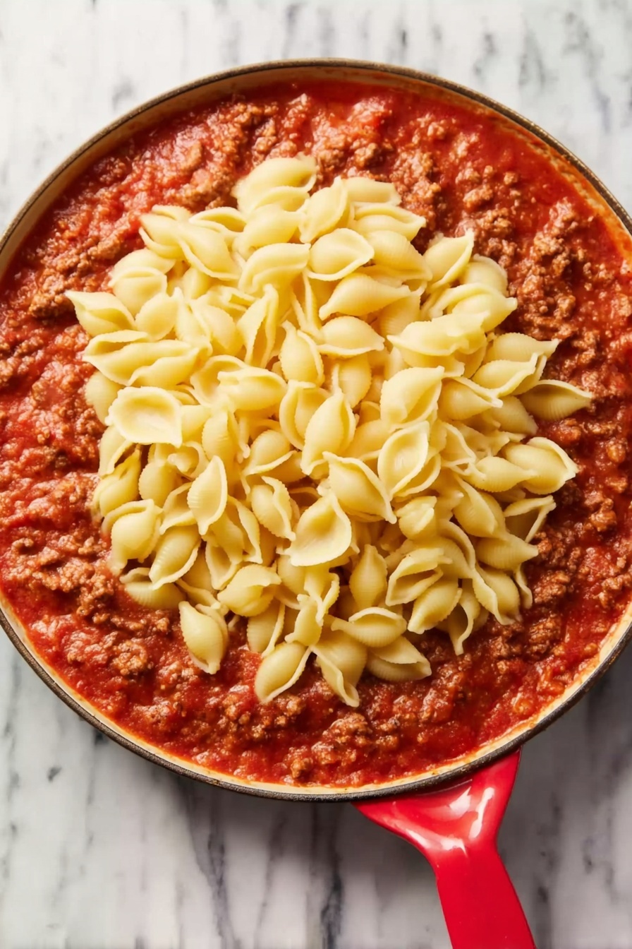 The image shows a close-up of a pasta dish with three main layers: the pasta shells, the meat sauce, and herbs. The pasta shells are golden yellow with ribbed texture and are the base layer. Mixed evenly with the shells is a thick, brown meat sauce containing small chunks of meat and soft onion pieces, making the dish look hearty and rich. Scattered on top and throughout are small green herb pieces that add a fresh touch of color. All layers are blended well together on a white marbled texture background. photo taken with an iphone --ar 2:3 --v 7